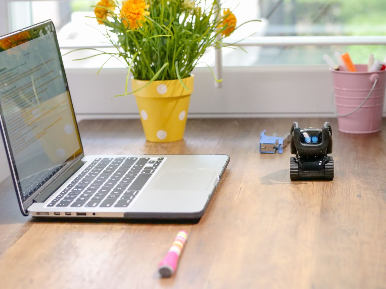 about-us-01 Stylish workspace featuring a laptop, toy robot, and vibrant flowers in natural light.